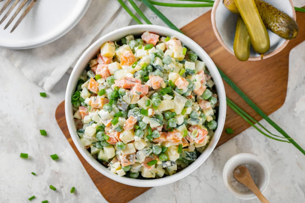 olivier-salad-in-a-white-bowl-on-a-wooden-board-with-green-onion-and-a-bowl-of-pickles-on-the-side