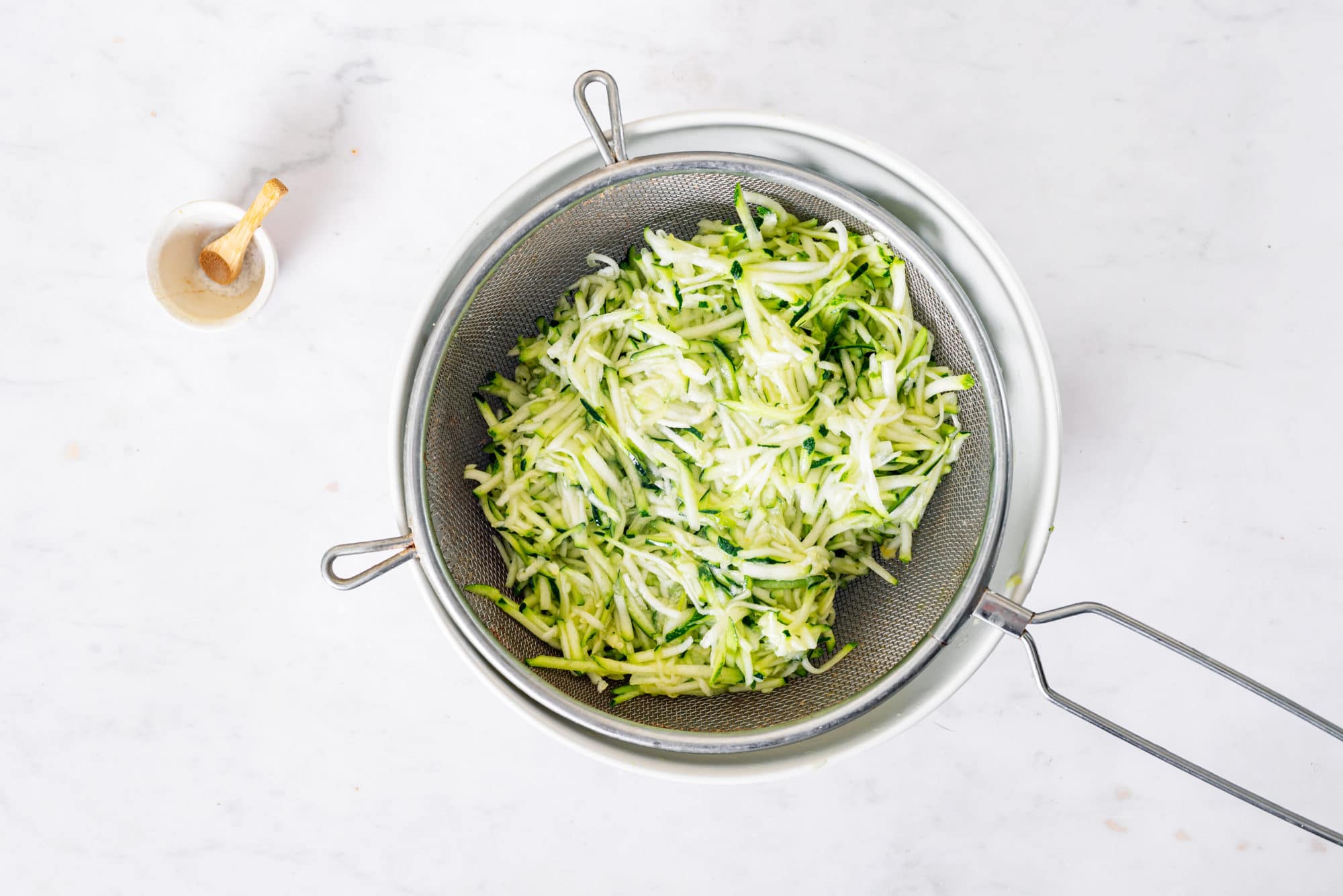 zucchini-shredded-in-a-sieve-over-a-white-bowl-with-salt-on-the-side