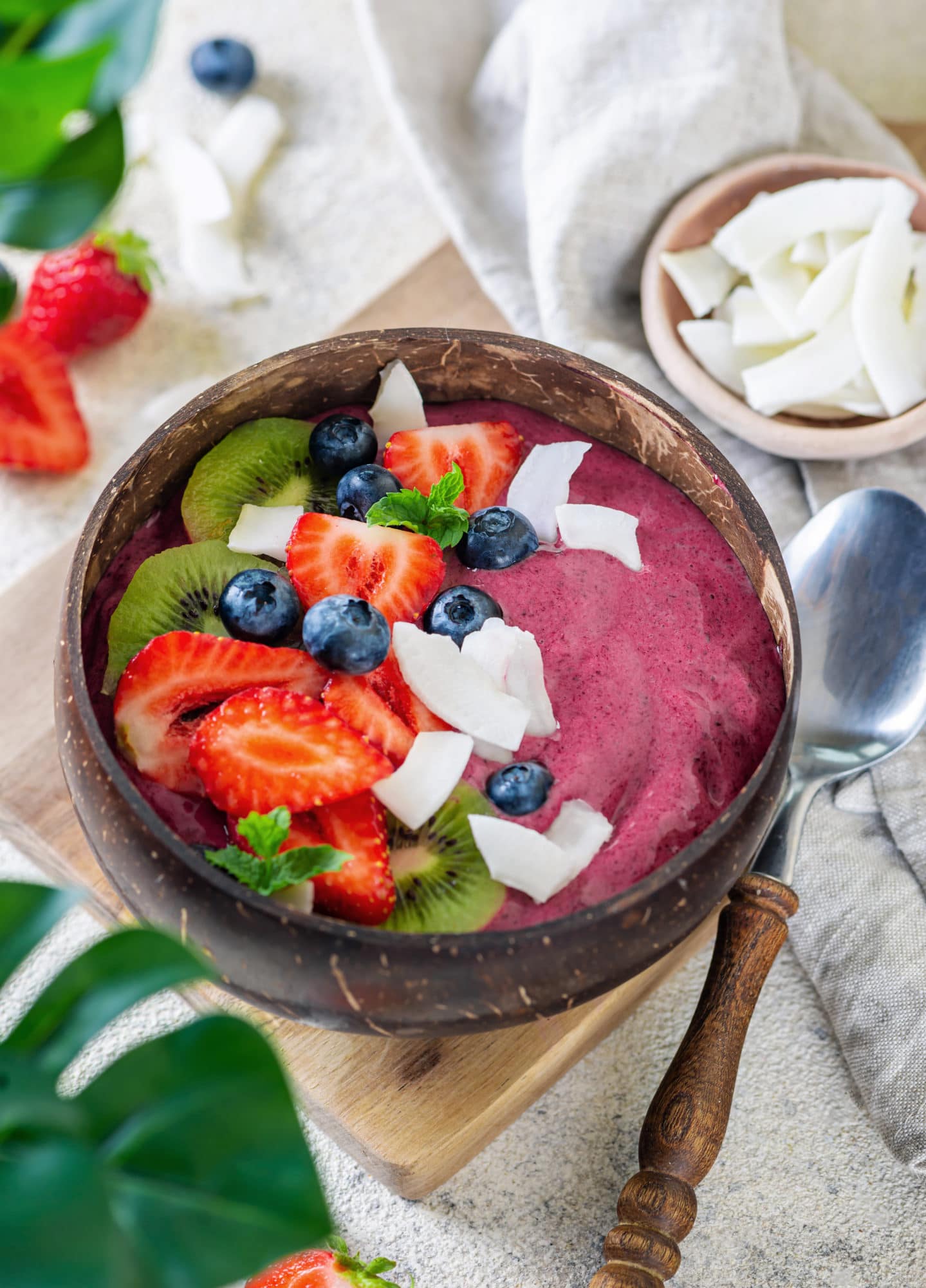 Acai smoothie in a brown coconut bowl topped with sliced strawberries, kiwi, blueberries, coconut slices and fresh mint surrounded by ingredients and a spoon on the side.