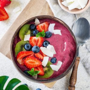 Acai smoothie in a brown bowl with toppings and a spoon, berries and coconut pieces on the side.