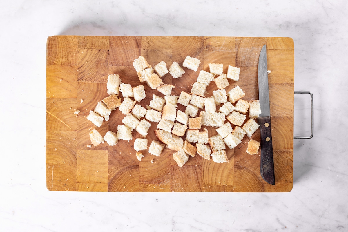 cut up bread into cube pieces on a wooden cutting board with small knife.