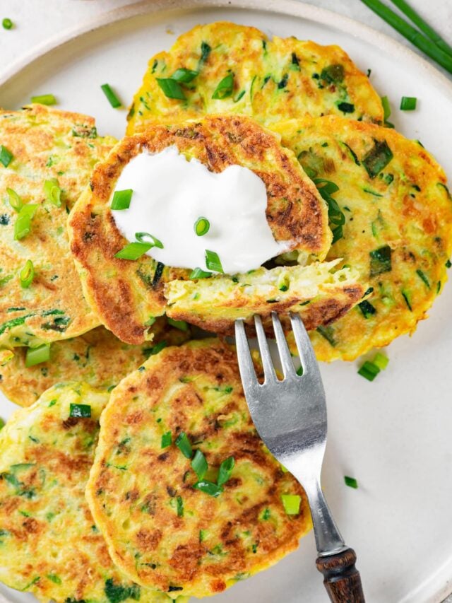zucchini fritters on a white plate with a fork sour cream and green onion on top.