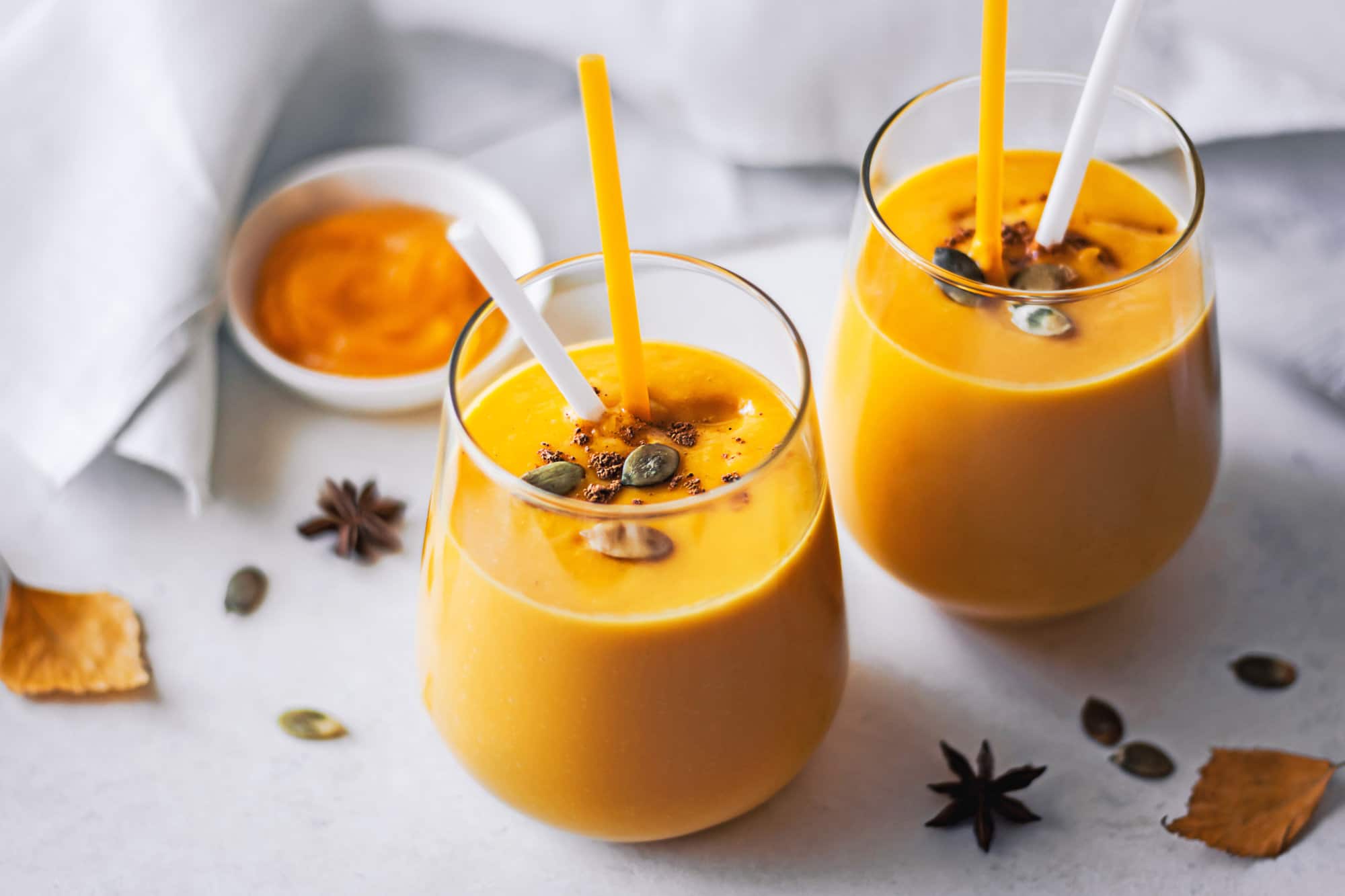 Pumpkin smoothie in a glass cup with a straw and topped with pumpkin seeds and spices.