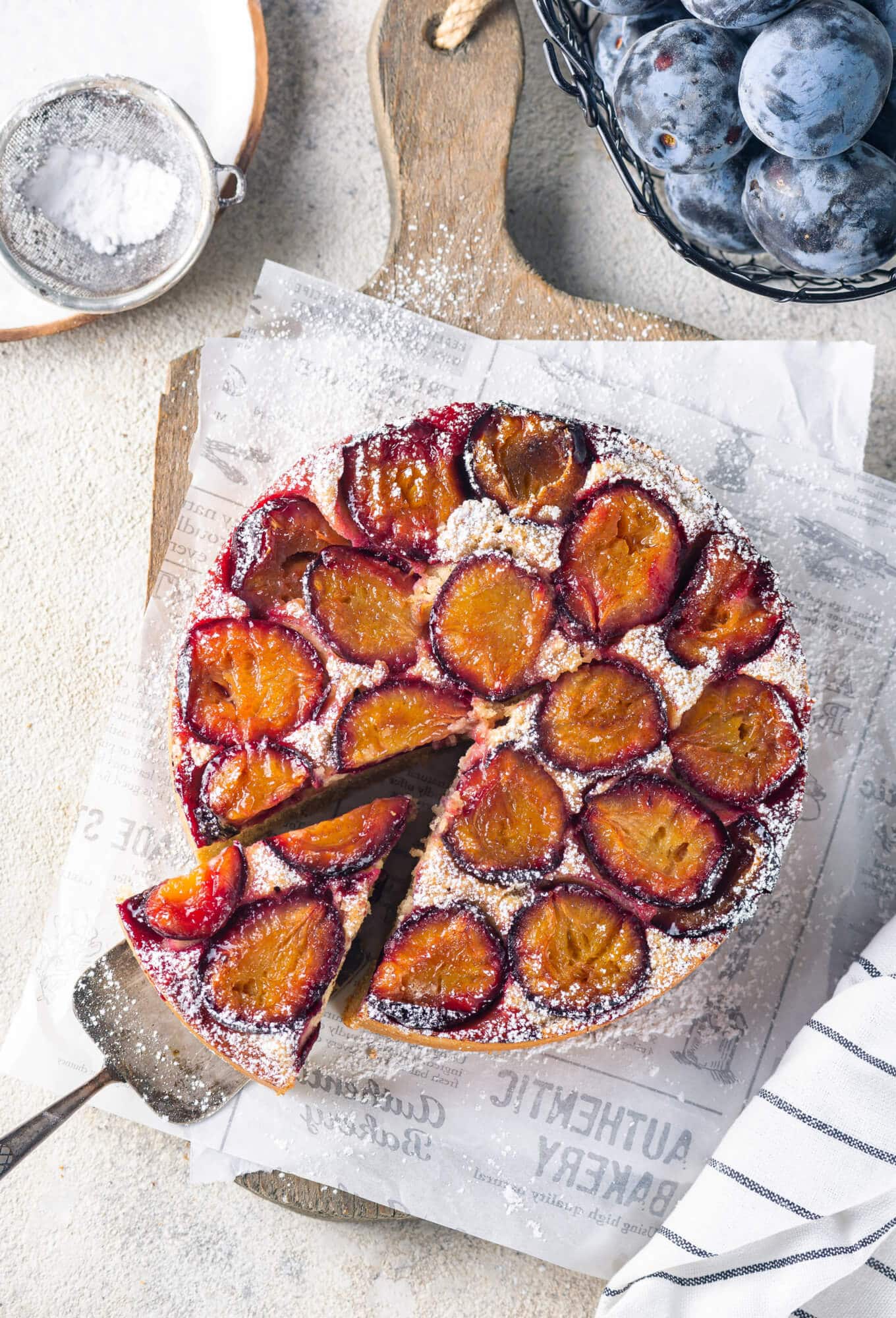 Plum cake atop white paper and a wooden board, with a spatula on the side for serving, and powdered sugar sprinkled on top.