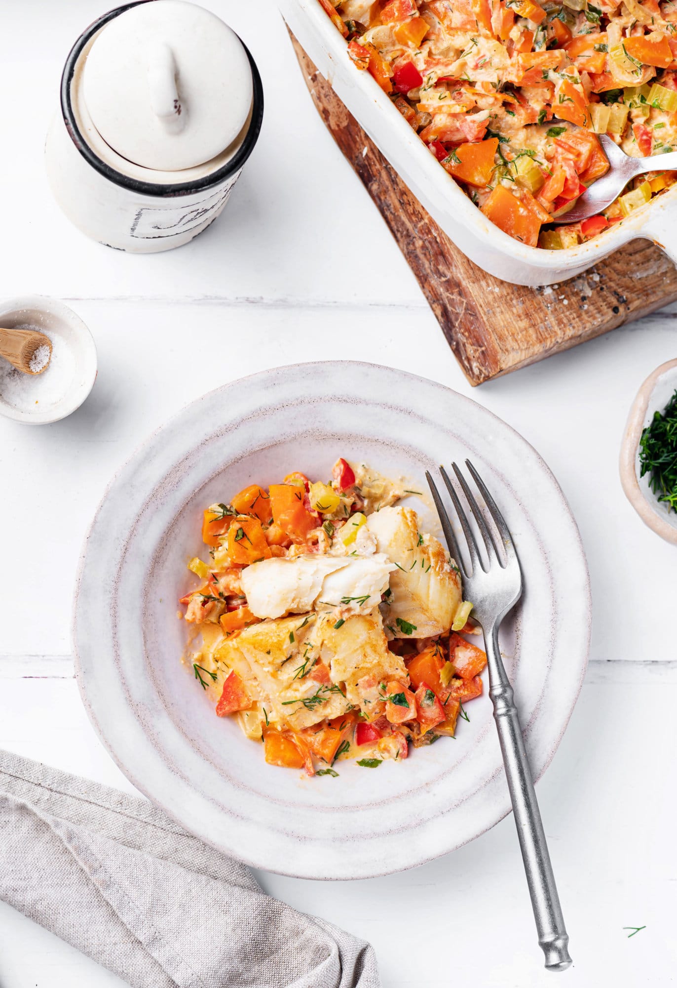 Fish and veggies in a bowl with a fork and a baking dish filled with more fish and veggies on the side.