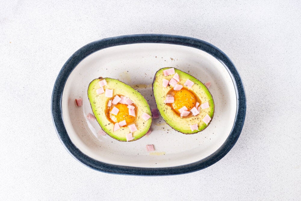 Raw avocado sliced in half with a raw egg in each avocado topped with bacon bits in a white baking dish.