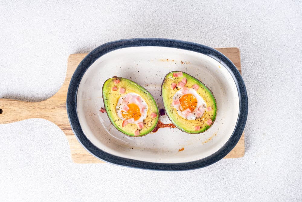 Baked avocado with egg in it and topped with bacon bits and green onion in a white baking dish.