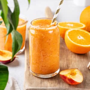 apricot smoothie in a glass cup with a straw and more halved oranges on the side.