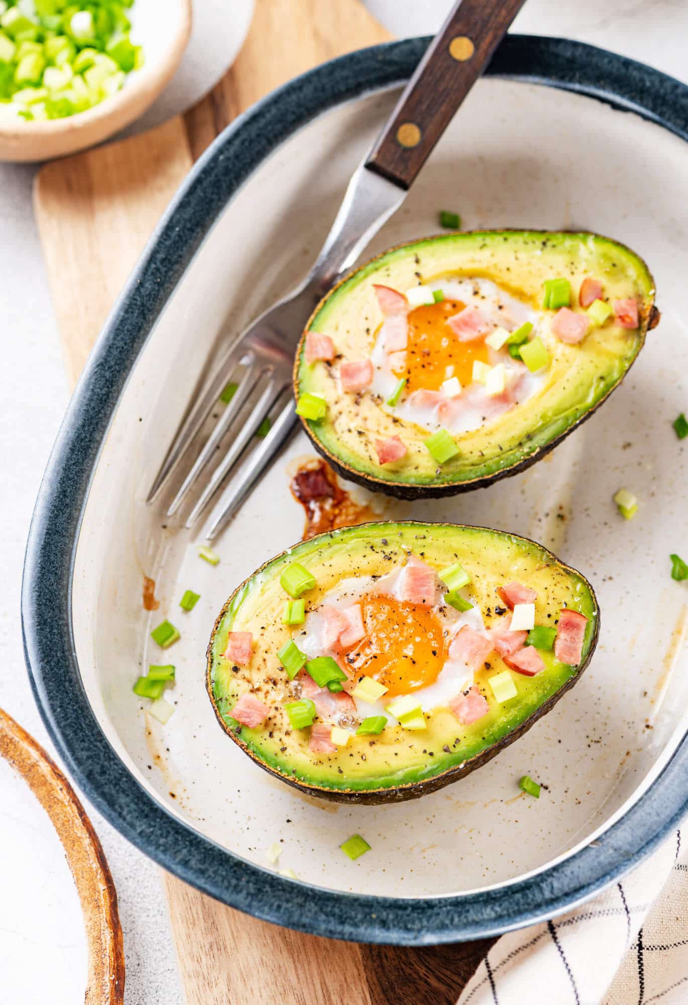 Baked avocado with egg in it and topped with bacon bits and green onion in a white baking dish with a silver fork on the side.