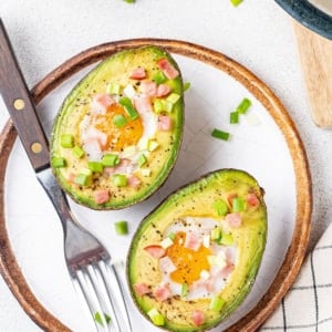 Baked avocado with egg in it and topped with bacon bits and green onion on a white plate with a silver fork on the side.