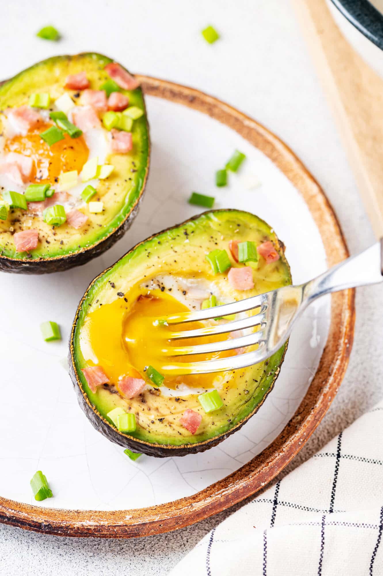 Baked avocado with an egg inside, topped with bacon and green onion on a white plate with a silver fork.