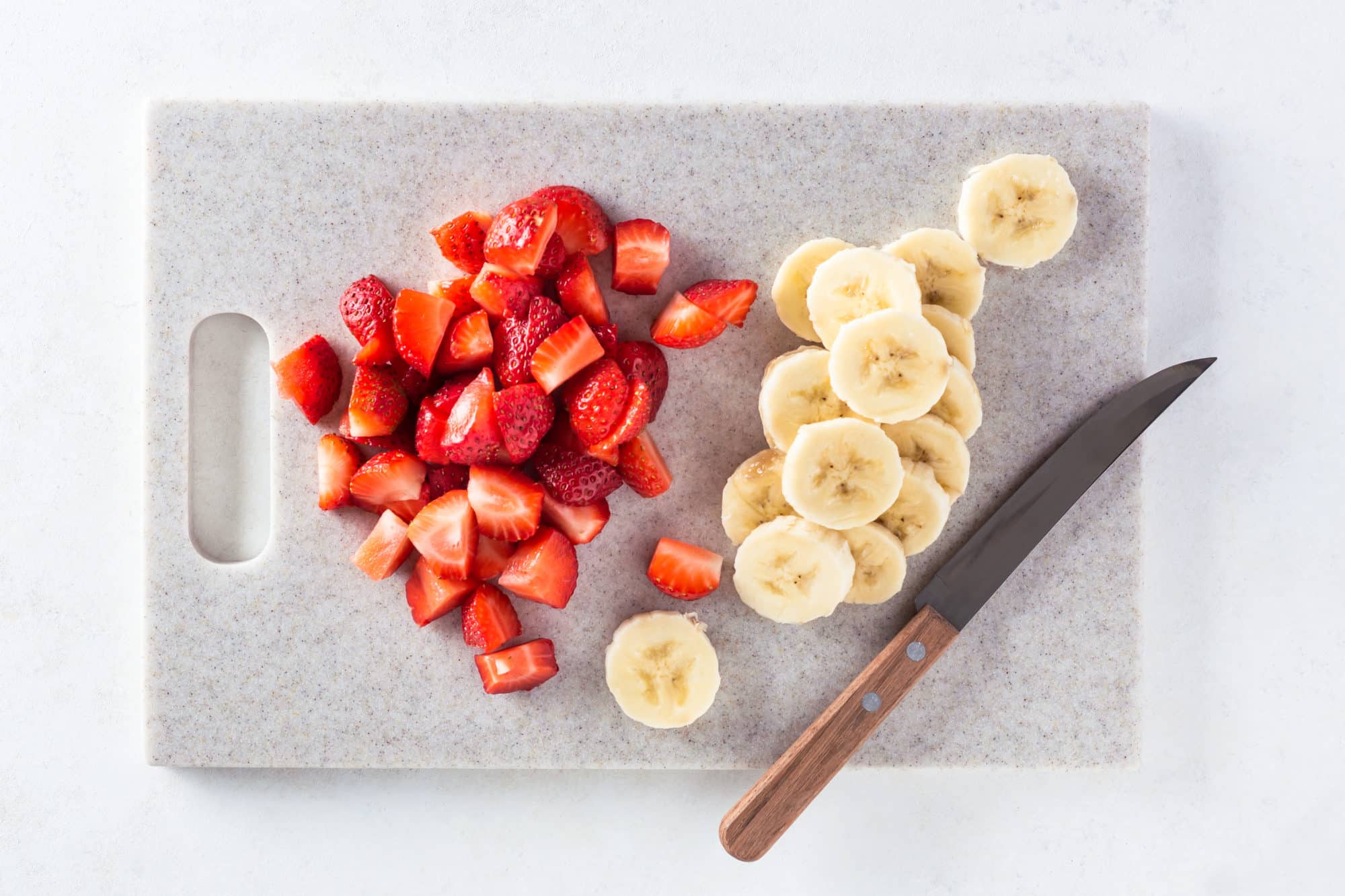 A cutting board with sliced strawberries and bananas and a knife. 
