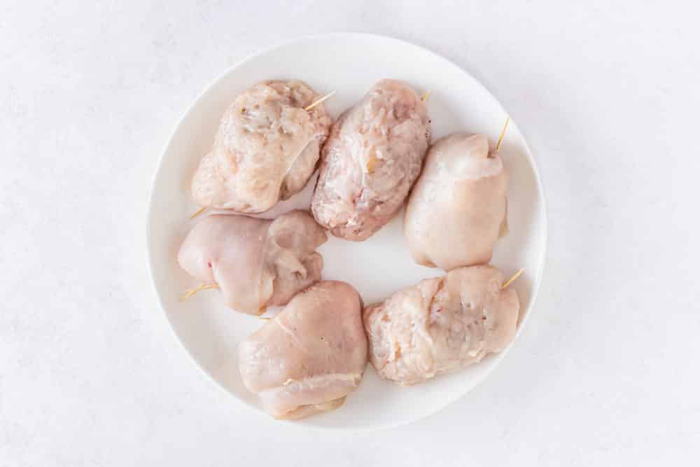 a white plate with raw rolled chicken breasts with toothpicks.
