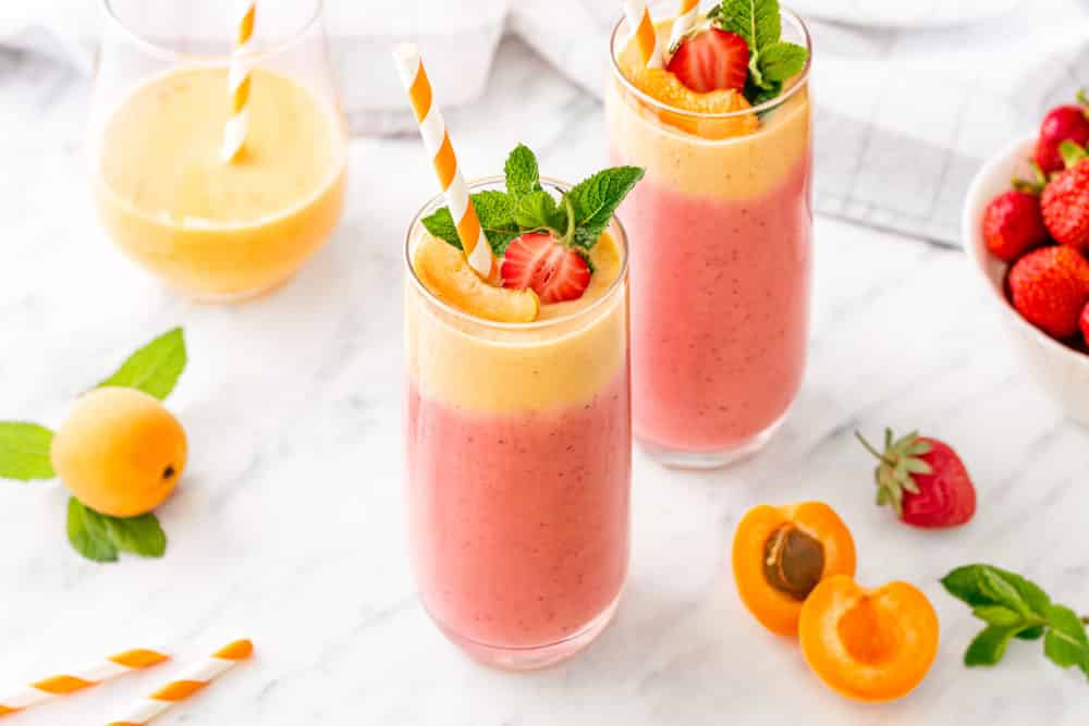 Apricot smoothie in a tall clear glass with a striped straw topped with fresh mint and half a strawberry surrounded by another glass of smoothie and fresh fruit.