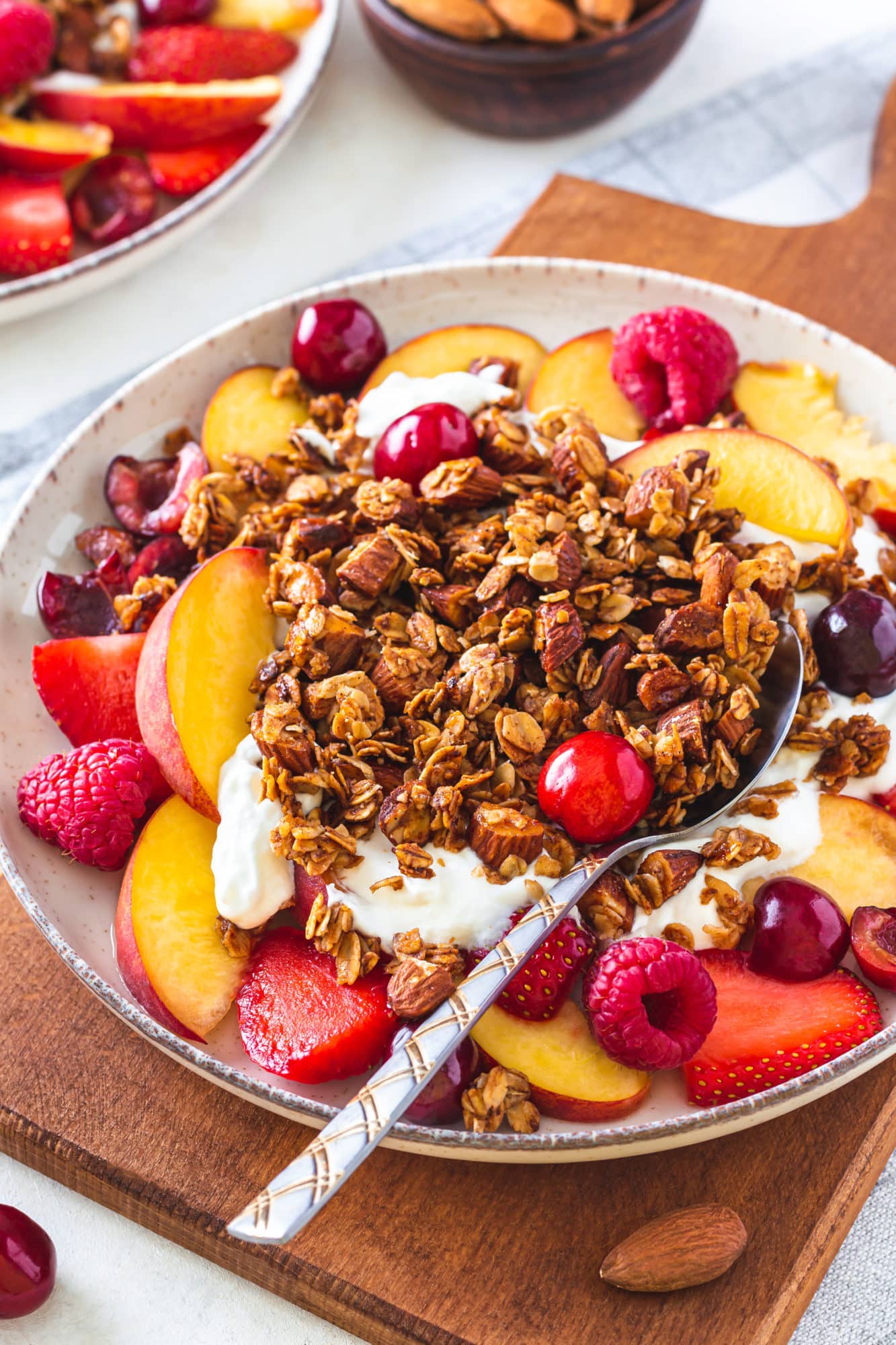 A bowl of muesli with yogurt, fruit and a spoon on a wooden cutting board. 