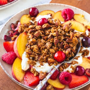 A bowl of muesli with yogurt, fruit and a spoon on a wooden cutting board.