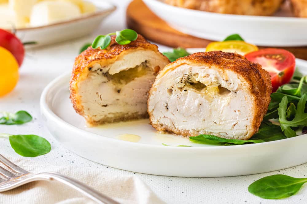 cooked butter stuffed chicken breast cut in half on a white plate with greens and tomatoes.