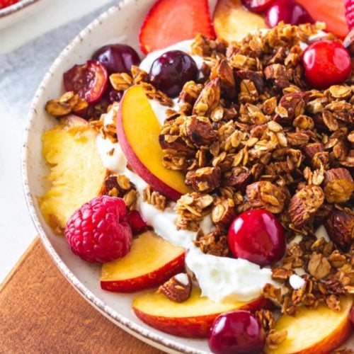 A bowl of muesli with yogurt and fruit on a wooden cutting board.