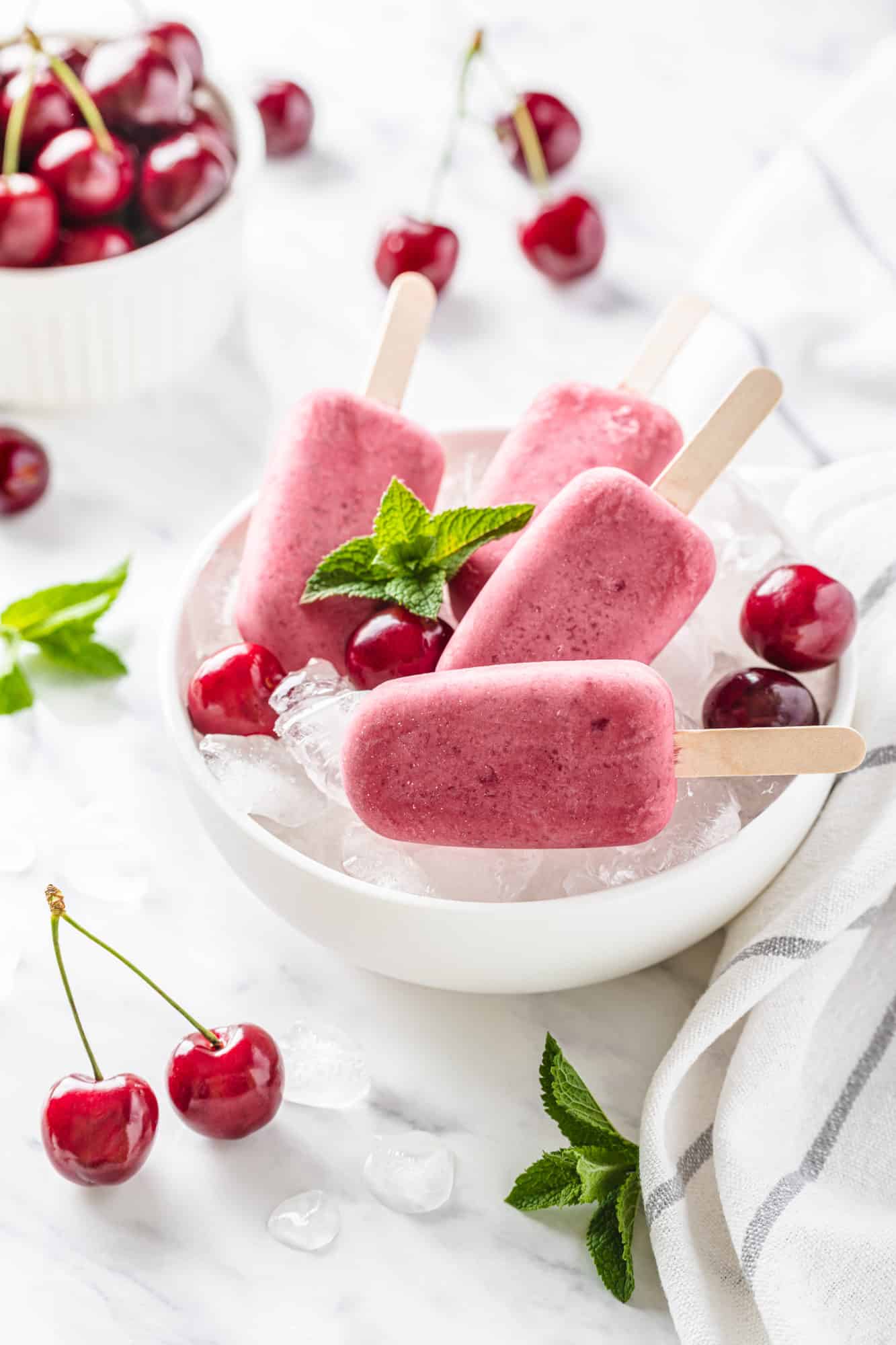Cherry ice pops in a white bowl filled with ice with cherries and mint all around.