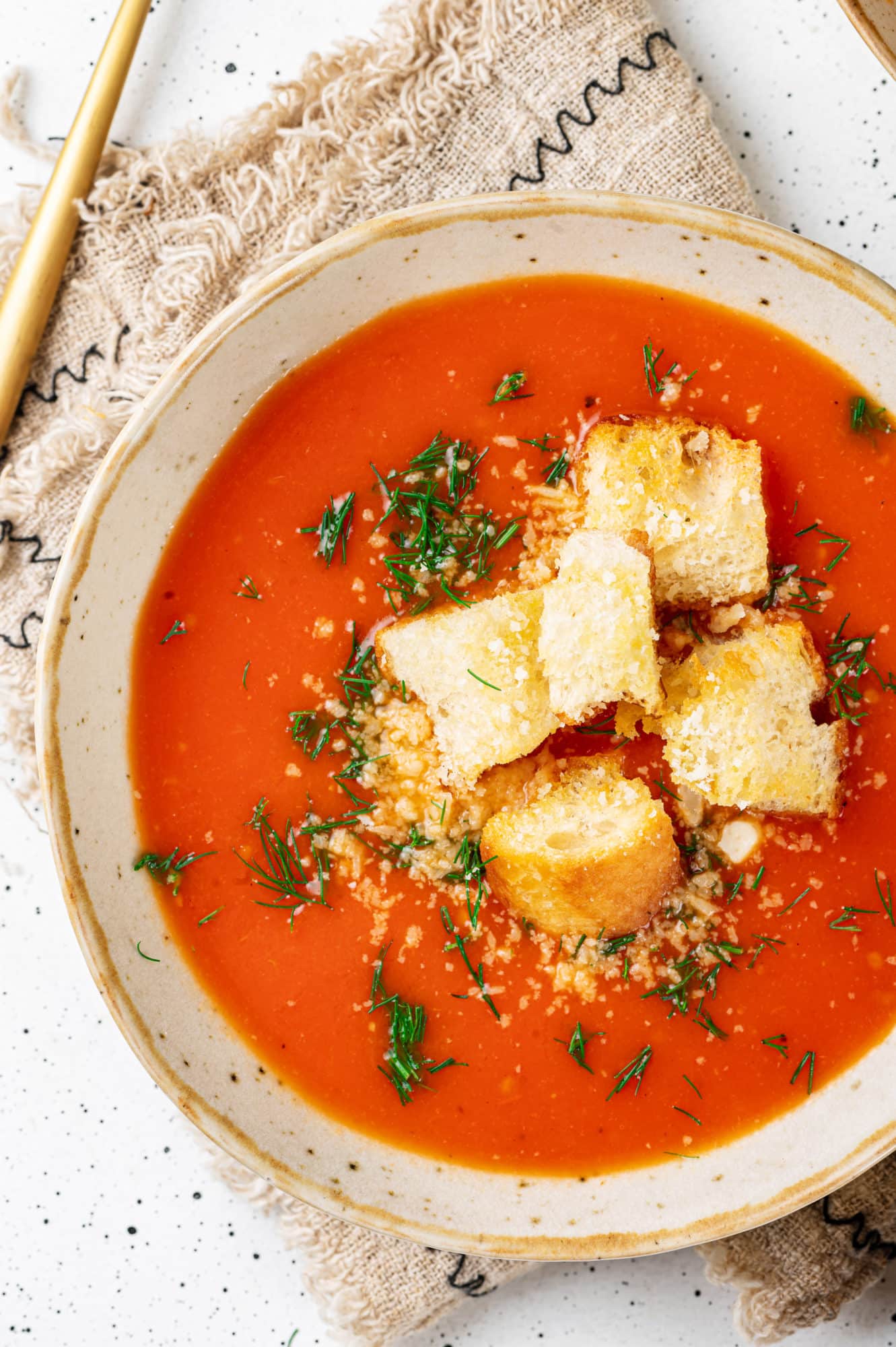 a beige bowl with red tomato soup and croutons and greens in it.