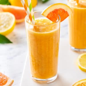 a glass filled with a citrus smoothie with two striped straws and a citrus wedge on the rim.