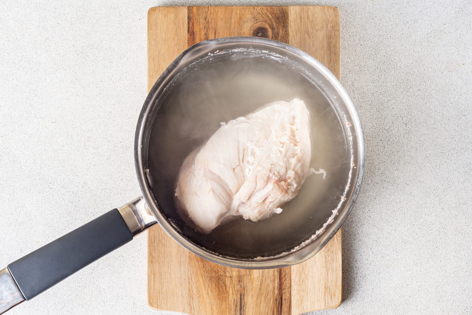 a chicken breast in a saucepan with water on a wooden cutting board.