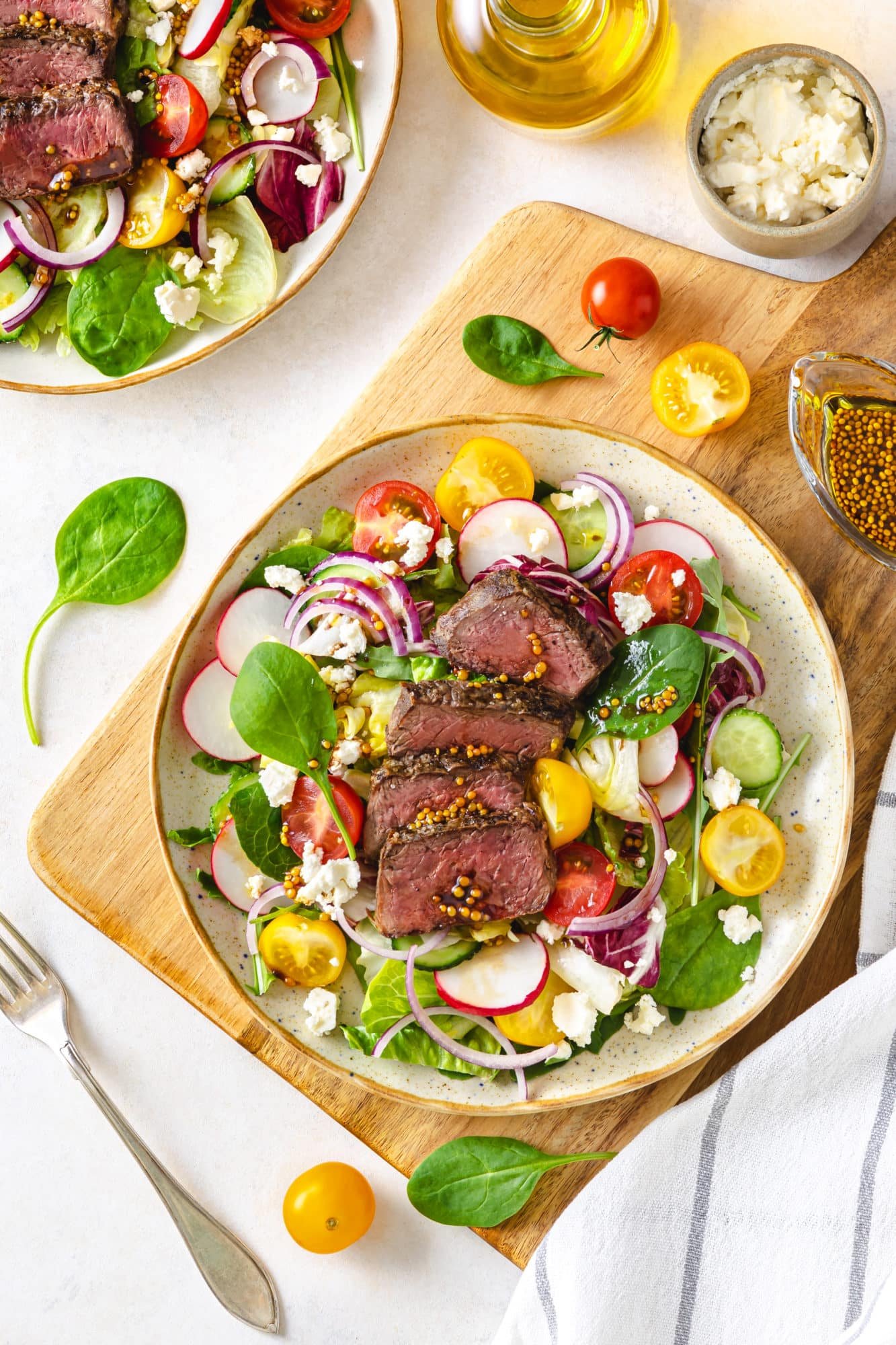 filet-mignon-salad-on-a-white-plate-on-a-wooden-nboard-with-tomatoes-and-greens-all-around-with-a-fork-on-the-side