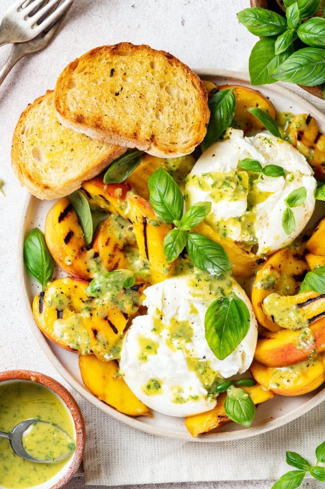 a plate with burrata peaches dressing and basil and two toasted bread slices with a bowl of dressing on the side.