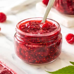 raspberry-jam-in-a-clear-jar-with-a-spoon-and-with-raspberries-and-more-jam-jars-in-the-background