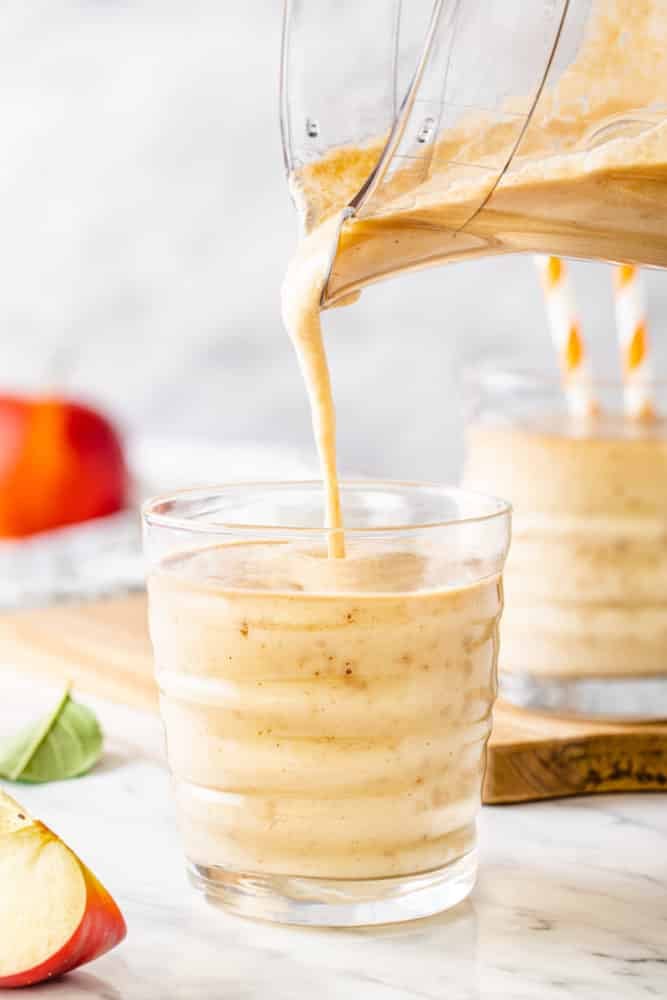 apple-smoothie-in-a-glass-with-smoothie-being-poured-from-a-glass-pitcher-with-a-wooden-board-in-the-background