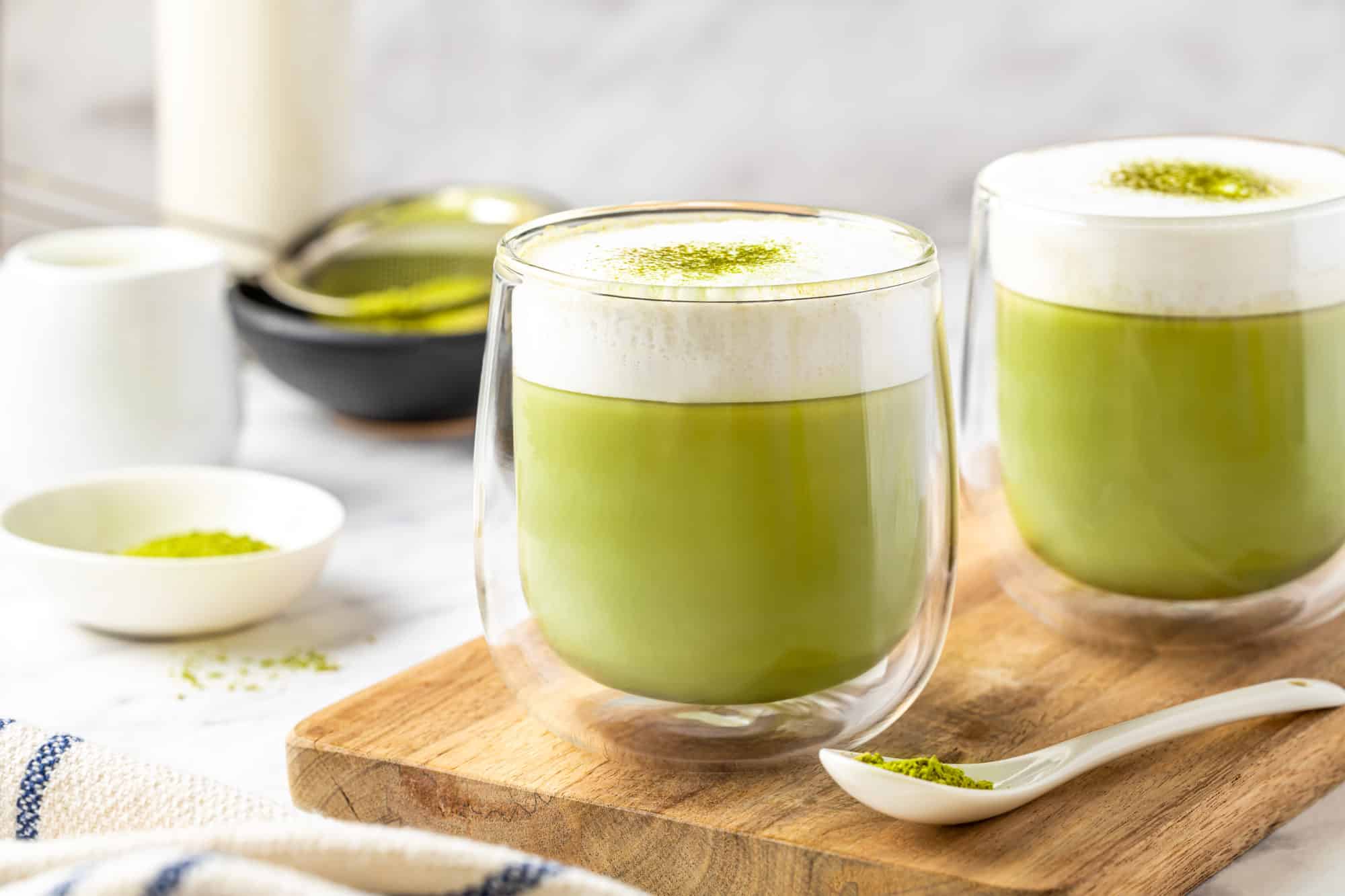 two glasses on a wooden board filled with matcha latte and frothed milk on top.