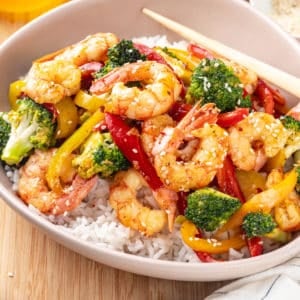 shrimp stir fry over rice in a dish on a wooden board with chopsticks on the side.