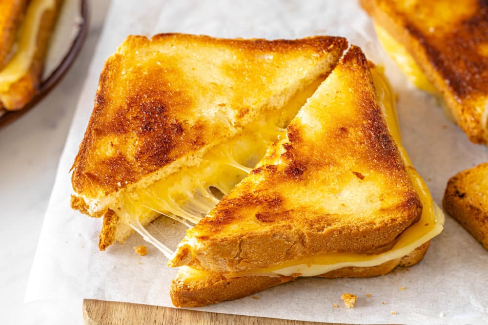 golden brown grilled cheese sandwich cut in half diagonally on parchment paper on a wooden board with cheese melting inside.
