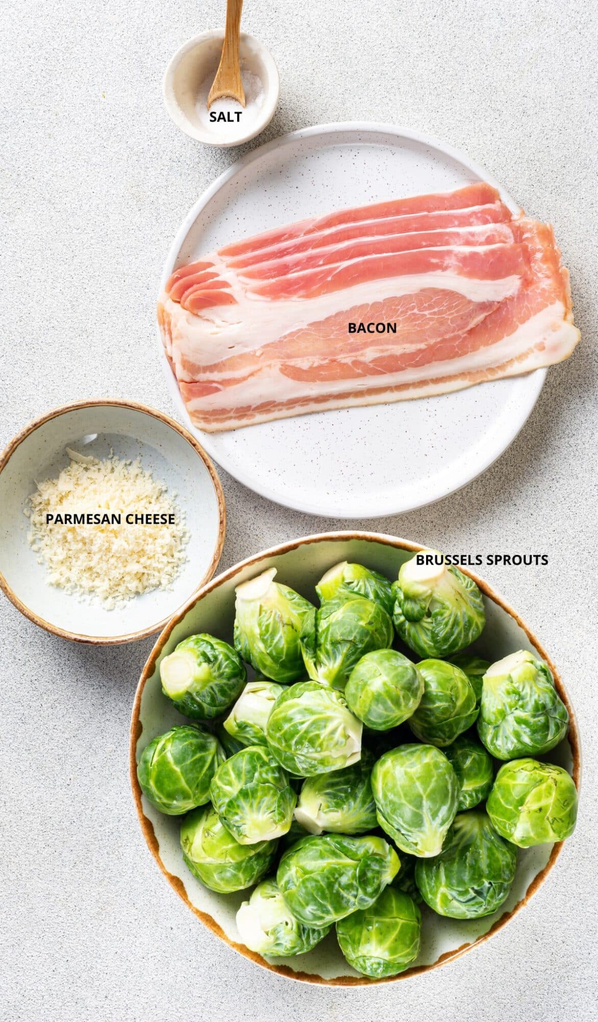 Brussels Sprouts in a bowl with grated parmesan cheese in another small bowl and raw bacon slices on a plate with salt and wooden spoon on the side.