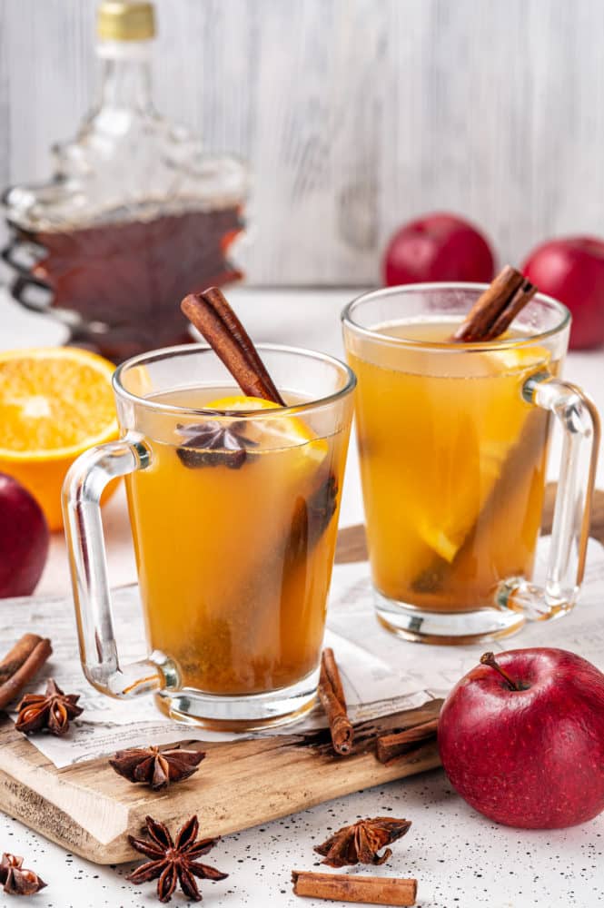 two glass mugs with apple cider and spices inside on a wooden baord with more cinnamon and star anise scattered around.