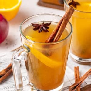 a wooden board with two mugs on it filled with apple cider and with spices floating inside.