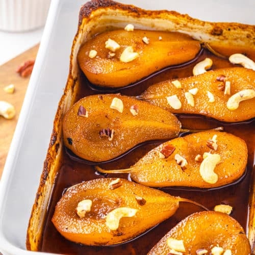 a wooden board with a baking tray on it filled with baked pears honey spice mix and cashews.