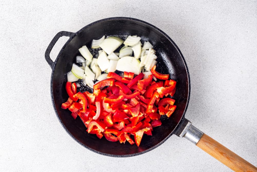 red bell peppers and onions sauteing in a black skillet with a wooden handle.