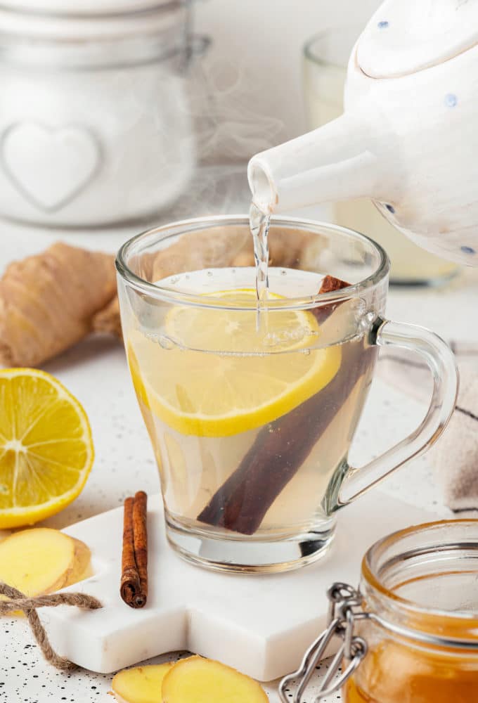 hot water being poured into a glass mug with ginger tea in it with a lemon slice and cinnamon stick floating in the tea on a white board with a cinnamon stick.
