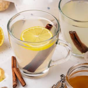 glass mugs filled with ginger tea with more flavor additions floating in the tea a lemon slice and cinnamon sticks.