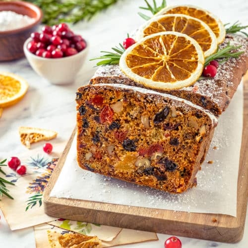fruit cake on a wooden board with parchment paper and one slice cut from the cake topped with powdered sugar candied oranges rosemary and cranberries with more toppings on the side.