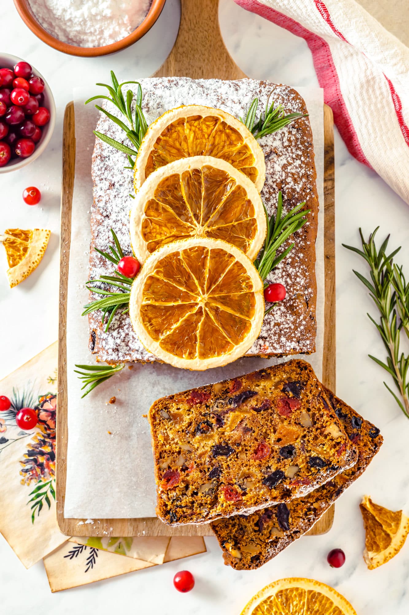 top view of a sliced fruit cake with toppings on top of the cake and more on the side including cranberries rosemary sprigs and some slices of candied orange.