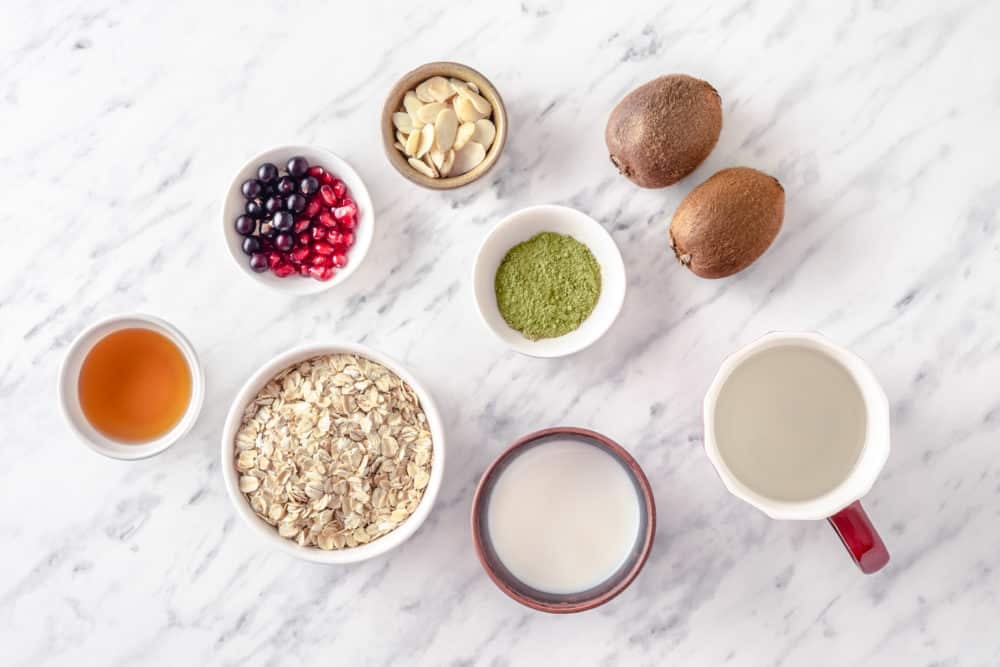 ingredients spread for a matcha oatmeal recipe in separate containers rolled oats milk water matcha powder maple syrup kiwis berries and almonds.