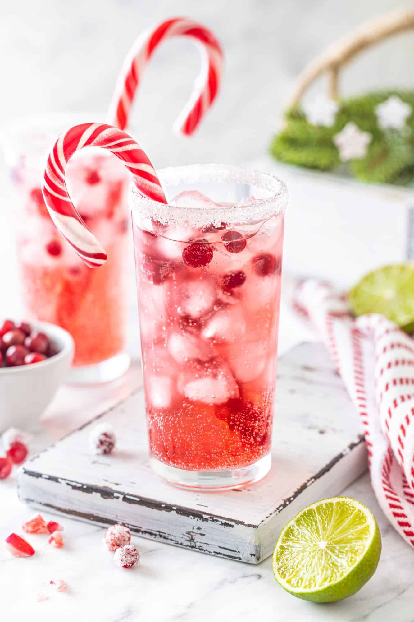 a pink and red mocktail drink put together in a glass cup with sugar on the rim and a candy cane in the cup on a wooden board with a lime wedge on the side.