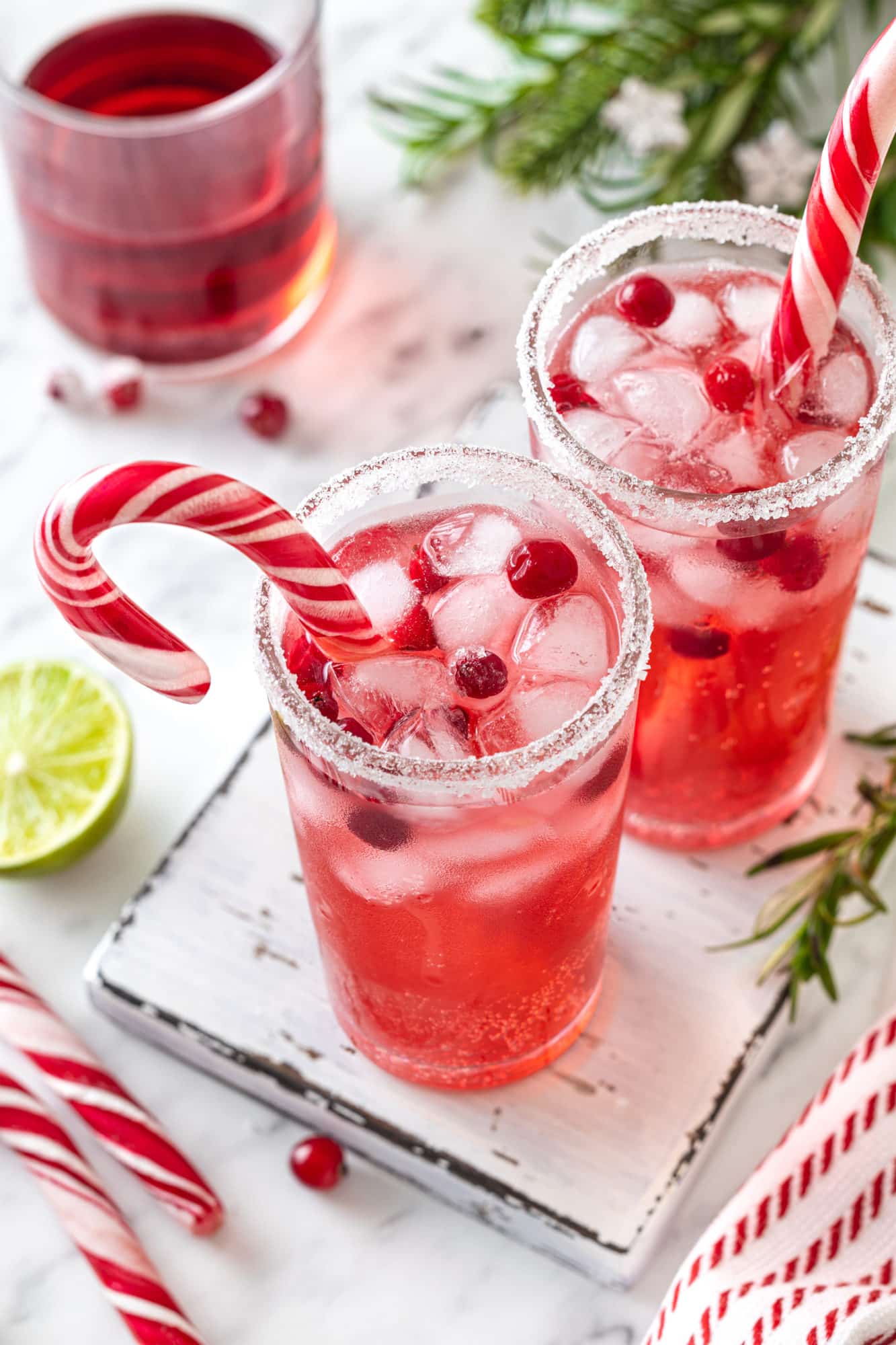 a peppermint drink in two glasses with sugar on the rim on a white wooden board with candy canes in the cups and christmas decorations scattered around.