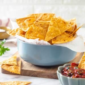 tortilla chips filling a blue bowl with parchment paper lining the bowl on a wooden board with more chips on the board and in a blue bowl filled with salsa.