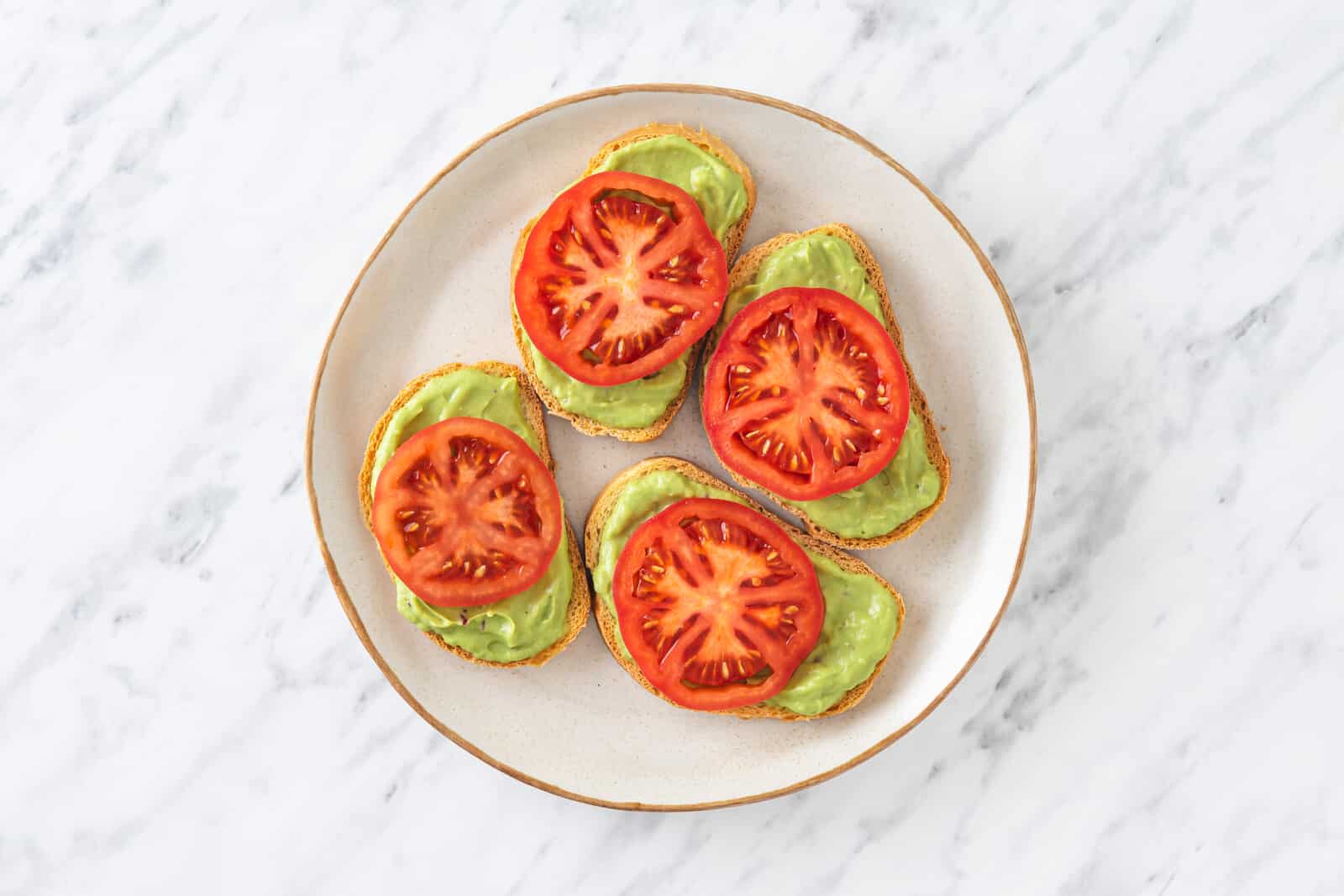 tomato slices on each avocado toast appetizer on a white plate.