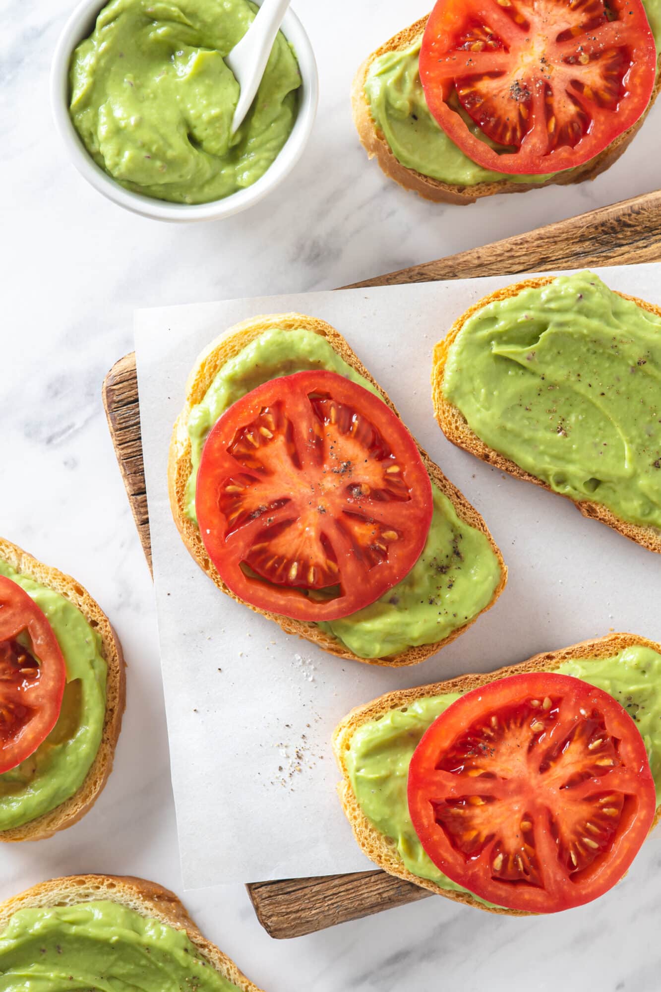 avocado toast pieces with a large tomato slice on each of them with some on a wooden board with parchment paper and a bowl of smooth avocado topping with a spoon in it.