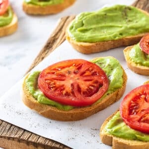 appetizers on parchment paper on a wooden board with a slice of tomato on each avocado toast.