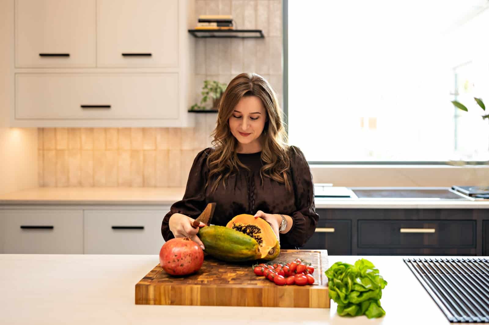 marina cutting open a papaya on a wooden board with a pomegranate cherry tomatoes and greens on the side in her kitchen.